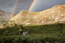Rückansicht einer Frau, die beim Wandern mit Hund im Urlaub den doppelten Regenbogen betrachtet — Stockfoto