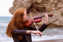 Junge rothaarige Geigerin spielt an einem bewölkten Tag ihre Geige am Strand — Stockfoto
