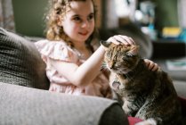Маленька дівчинка грає зі своїм котом на дивані у вітальні — стокове фото