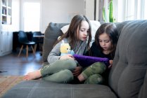 Дві маленькі дівчата сидять на дивані за допомогою планшета . — стокове фото