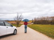 Donna con ombrello in mezzo alla strada accanto al campo con auto — Foto stock