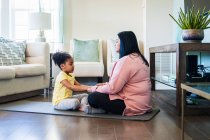 Abuela practicando meditación con su nieta mientras está sentada en la alfombra de ejercicio en la sala de estar en casa - foto de stock