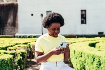 Портрет черной девушки с афроволосами и кольцевыми серьгами с помощью мобильного телефона в городском пространстве города. — стоковое фото