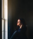 Mulher sentada sozinha em um quarto escuro olhando pela janela. — Fotografia de Stock