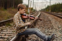 Un bel ragazzo biondo siede sulle rotaie della ferrovia e interpreta il v — Foto stock