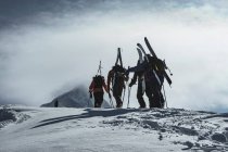 Rear view of people with splitboards and backpacks walking on snow covered mountain peak against cloudy sky — Stock Photo