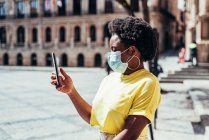 Porträt eines schwarzen Mädchens mit Gesichtsmaske, Afrohaaren und Reifrohren, das sein Handy auf einem alten Platz benutzt. — Stockfoto
