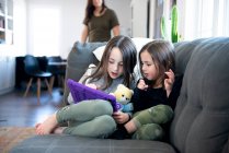 Two little girls sitting on the couch using a tablet. — Stock Photo