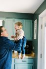 Pai segurando seu menino no ar de pé em sua cozinha — Fotografia de Stock