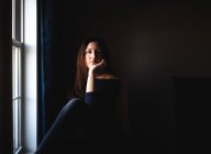Mulher atraente sentada sozinha em um quarto escuro ao lado de uma janela. — Fotografia de Stock