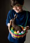 Jeune garçon heureux tenant panier de Pâques plein d'œufs et lapin en chocolat — Photo de stock