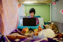 Мальчик сидит в постели в вязаной шляпе и делает домашнюю работу на компьютере с котом — стоковое фото