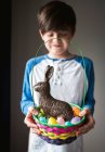 Jeune garçon heureux tenant panier de Pâques plein d'œufs et lapin en chocolat — Photo de stock