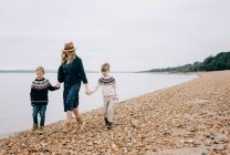 Mamá caminando por la playa cogida de la mano con sus hijos sonriendo - foto de stock