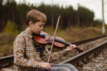 Ein schöner blonder Junge sitzt auf den Schienen der Eisenbahn und spielt den V — Stockfoto