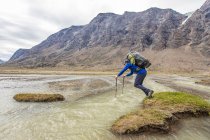 Backpacker utiliza bastones de trekking para saltar a través de un canal de río profundo - foto de stock