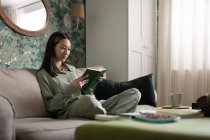 Jovem asiática relaxante no sofá e livro de leitura no fim de semana em casa — Fotografia de Stock