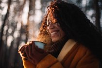 Щаслива молода жінка тримає чашку чаю, що стоїть в лісі взимку — стокове фото