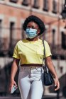 Chica afroamericana con máscara facial caminando por una calle de la ciudad. - foto de stock