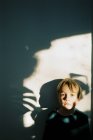 Petit garçon avec des ombres sur le visage et le mur derrière lui — Photo de stock