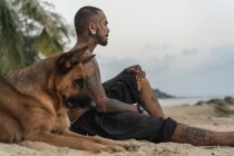 Asiático chico sentado en la playa con un perro entre las palmeras - foto de stock