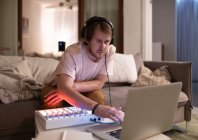 Дорослий чоловічий музикант переглядає ноутбук біля звукової дошки, сидячи на дивані вдома — стокове фото