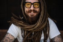 Портрет хлопця-хіпстера в окулярах і з дредлоками і татуюванням сидить на тлі дерев'яного будинку — стокове фото