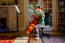 Босоногий мальчик практикует виолончель в помещении с золотым окном — стоковое фото