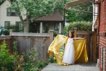A little girl stands in garden arms outstretched in long gold cape — Stock Photo
