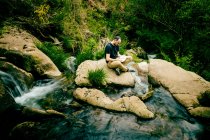 Hombre leyendo un libro sentado en una roca junto a un río - foto de stock