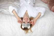 Young attractive smiling woman in sleep mask and bathrobe lie on bed, looking at camera — Stock Photo