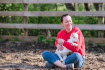 Woman in red sweatshirt holding a white baby goat on her lap. — Stock Photo