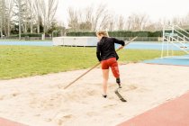 Back view of female athlete with prosthetic leg using pole to prepare sand for jump during track and field workout on stadium — Stock Photo