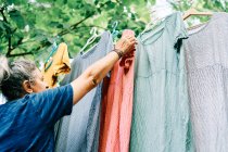 Woman hangs colorful dresses on hangers on clothesline in home garden — Stock Photo