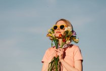 Mujer de estilo en vestido rosa con gafas amarillas sosteniendo flores sobre fondo azul cielo - foto de stock