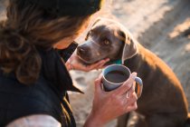 Молода жінка зі своїм собакою п'є каву на пляжі — стокове фото