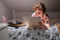Mujer joven acostada en su cama, escuchando un disco de vinilo y mirando a la manga - foto de stock