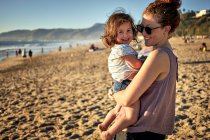 Seitenansicht einer glücklichen Mutter, die ihre Tochter am Wochenende am Strand trägt — Stockfoto