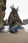 Homem sentado na praia com pilhas de mar vestindo um casaco inchado e chapéu — Fotografia de Stock