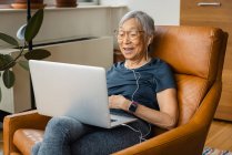 Porträt einer Seniorin, die Kopfhörer trägt, während sie zu Hause ihren Laptop benutzt — Stockfoto