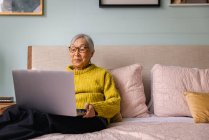 Donna anziana che utilizza il computer portatile mentre seduto in camera da letto a casa — Foto stock