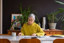 Mujer mayor con suéter amarillo leyendo el periódico mientras está sentada en la mesa de comedor - foto de stock