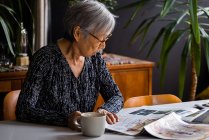 Mujer mayor leyendo el periódico mientras está sentada en la mesa con café - foto de stock