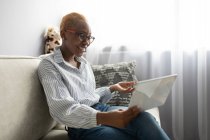 Positive junge Afroamerikanerin in lässigem Outfit und Brille sitzt auf dem Sofa und kommuniziert mit Geschäftspartnern per Videoanruf auf dem Tablet, während sie von zu Hause aus fernarbeitet — Stockfoto