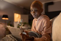 Joven mujer afroamericana en sudadera con capucha informal bebiendo vino y viendo películas en tabletas mientras se relaja en un acogedor sofá en la sala de estar por la noche - foto de stock