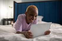 Улыбающаяся молодая афроамериканка лежит на кровати и занимается серфингом в Интернете на планшете, проводя свободное время дома — стоковое фото