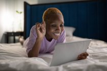 Молодая черная женщина наслаждается онлайн видео-чатом с другом, лежа на кровати и отдыхая в свободное время дома — стоковое фото