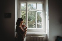 Мати тримає новонароджену дитину в кімнаті, дивлячись у вікно — стокове фото