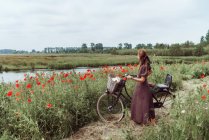 Donna con bicicletta in piedi tra papaveri campo contro il cielo — Foto stock
