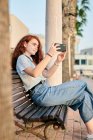 Молода руда жінка робить фотографію зі своїм мобільним телефоном, сидячи в лавці — стокове фото